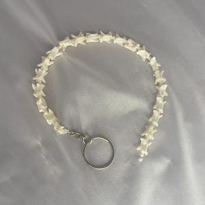 May include: A white, bone-like bracelet with a silver-coloured key ring. The bracelet is made of small, connected pieces, forming a curved shape. The background is a soft, white fabric.