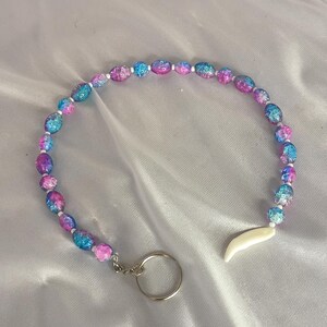 May include: A beaded keychain with a galaxy design. The oval beads are a mix of blue, pink, and purple. It has a silver keyring and a white, tooth-shaped charm attached. The beads are approximately 1 cm in diameter.
