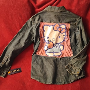 Puede incluir: Una camisa de mezclilla azul de manga larga con un parche de Hello Kitty en la espalda. El parche es rosa y presenta a Hello Kitty sosteniendo una manzana. La camisa tiene una etiqueta de Cherokee en la parte inferior izquierda.