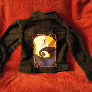 Könnte beinhalten: Schwarze Jeansjacke mit einem aufgenähten Patch, der Jack Skellington aus Nightmare Before Christmas zeigt. Der Patch zeigt Jack, der auf einem Hügel mit einem Halbmond im Hintergrund steht.