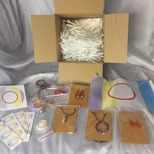 Op de afbeelding: Een open kartonnen doos gevuld met wit verpakkingsmateriaal, omringd door diverse sieraden en accessoires. De items omvatten kettingen, oorbellen, stickers en kleine vlaggen met een wit ovaal ontwerp en rode omtrek.