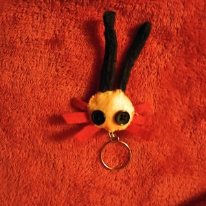 May include: A handmade keychain featuring a whimsical creature design. The creature has a cream-colored head with two large black button eyes, black antennae, and a red bow. A gold-colored keyring is attached to the bottom.