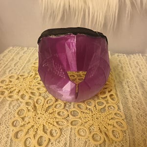 May include: A purple, faceted mask with a black top edge. The mask is on a cream-coloured, lace doily. A fluffy, white surface is in the background.