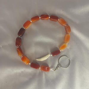 May include: A bracelet featuring rectangular, orange and brown beads strung together with white string. A small, white, curved charm and a silver-colored key ring are attached. The beads transition in color from dark brown to light orange.