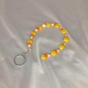 May include: A keychain with a string of candy corn-shaped beads in yellow, orange, and white. The beads are linked together and attached to a silver-coloured key ring. The candy corn design is a classic Halloween theme.
