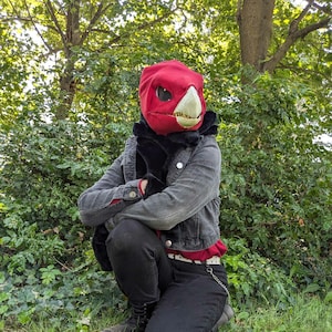 May include: A person wearing a red and white animal mask with a black jacket and black pants. The person is standing in a forest setting.