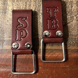May include: Two brown leather tool belt loops with metal D-rings. The loops are personalized with the initials "SP" and "TB" in a decorative font.