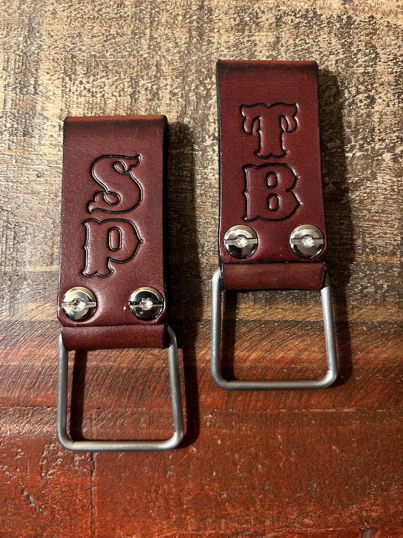 May include: Two brown leather tool belt loops with metal D-rings. The loops are personalized with the initials "SP" and "TB" in a decorative font.