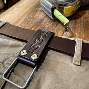 May include: A brown leather belt loop with a metal ring and the word "7even" embossed on it. The loop is attached to a brown leather belt and is sitting on a beige fabric surface. A silver tape measure is also visible in the background.