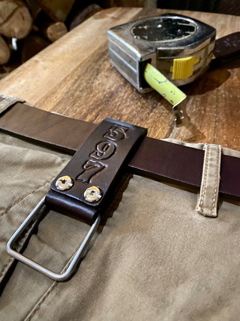 May include: A brown leather belt loop with a metal ring and the word "7even" embossed on it. The loop is attached to a brown leather belt and is sitting on a beige fabric surface. A silver tape measure is also visible in the background.