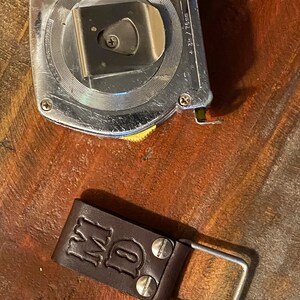 May include: A silver metal tape measure with a yellow knob and a brown leather belt loop with the initials "MD" embossed on it.