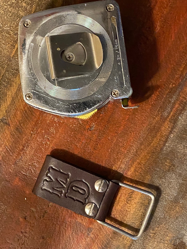 May include: A silver metal tape measure with a yellow knob and a brown leather belt loop with the initials "MD" embossed on it.
