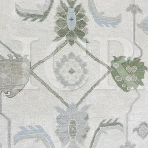 May include: Close-up of a cream-colored rug with a detailed floral pattern. The design features stylized flowers and geometric shapes in shades of green, blue, and gray. The rug has a textured appearance, suggesting a woven construction.