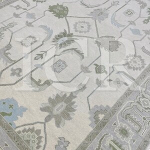May include: A detailed close-up of a beige area rug with an intricate floral and geometric pattern. The rug features muted tones of gray, green, and blue, creating a soft, inviting aesthetic. The design includes stylized flowers and repeating shapes.