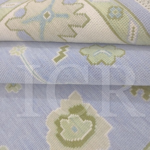 May include: Close-up of a folded area rug with a floral pattern. The rug features a light blue background with green, white, and light blue floral and geometric designs. The rug appears to be made of a soft, textured material.