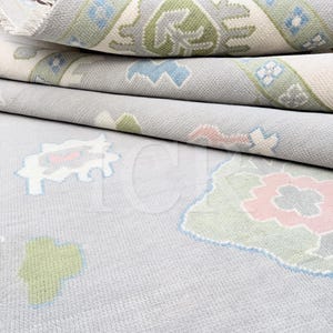 Peut inclure: Gros plan sur des tapis pliés avec un fond gris clair. Les tapis présentent des motifs complexes dans des tons de vert, de bleu, de rose et de blanc. Les motifs comprennent des formes florales et géométriques, créant un effet de superposition. Les bords des tapis sont visibles, mettant en valeur le savoir-faire.