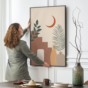 May include: Abstract art print featuring a terracotta-colored staircase, a crescent moon, and green foliage against a beige background. The artwork is framed in black and is being held by a person. The overall theme is modern and minimalist.