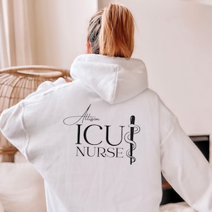 Puede incluir: Sudadera con capucha blanca con el texto "Alison ICU NURSE" y un símbolo de caduceo impreso en la espalda.