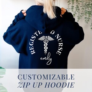 May include: A navy blue zip-up hoodie with a white design on the back. The design features a caduceus, the symbol of medicine, with the words "Registered Nurse" and a name, "emily".