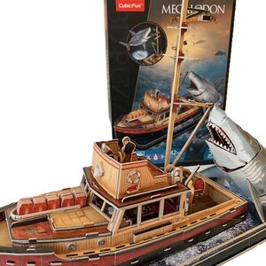 May include: A wooden boat puzzle with a shark figure. The boat is brown and red, with a mast and a small cabin. The puzzle box in the background features a shark and the word "Megalodon".