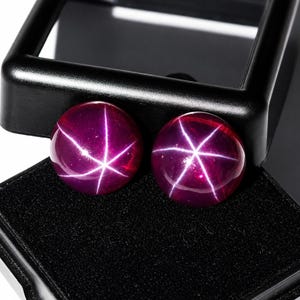 May include: Two round, deep magenta cabochon gemstones, each displaying a distinct six-rayed star pattern across its smooth, polished surface. The stones are positioned side-by-side, resting within a black display case with a velvet-like interior.