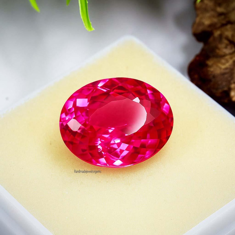 May include: A faceted, oval-cut, vibrant pink gemstone. The gem is set on a cream-colored surface. The image is well-lit, showcasing the gem's brilliance and clarity. The text "handmadewelgems" is visible.