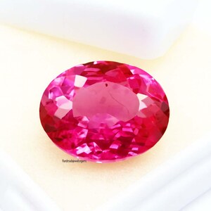 May include: A faceted, oval-shaped, hot pink gemstone. The gem is set against a white background, with the text "Handmadejewelgems" visible. The gemstone's facets reflect light, creating a sparkling effect. This gem is suitable for jewelry making.