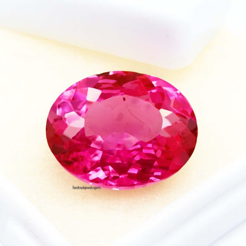 May include: A faceted, oval-shaped, hot pink gemstone. The gem is set against a white background, with the text "Handmadejewelgems" visible. The gemstone's facets reflect light, creating a sparkling effect. This gem is suitable for jewelry making.