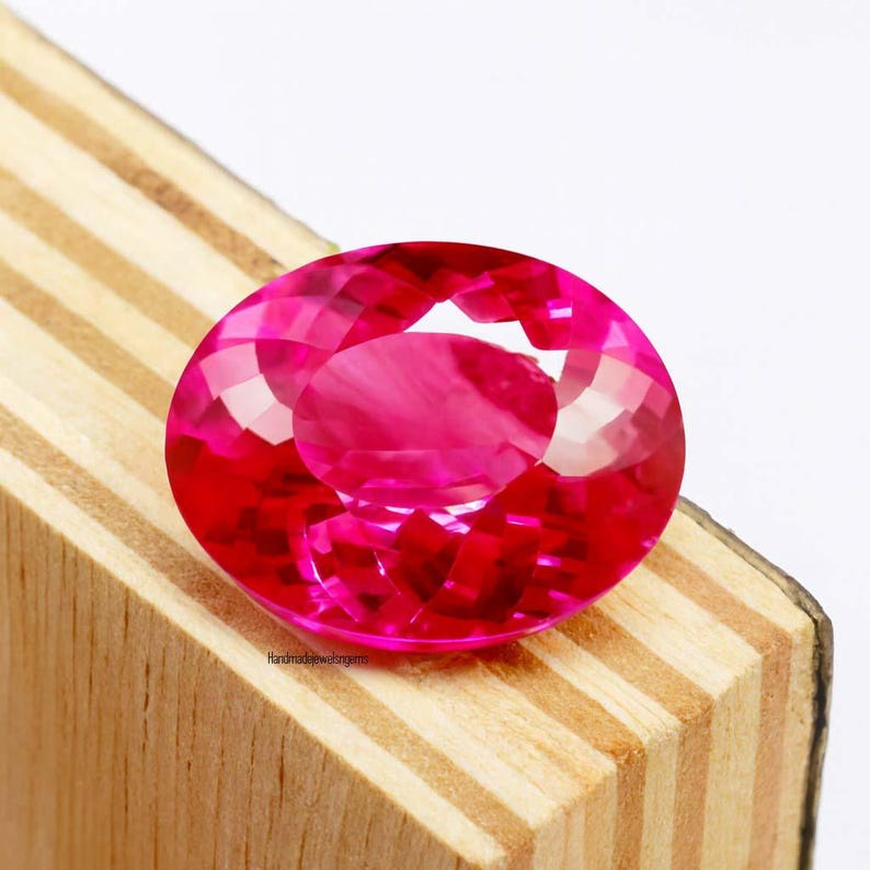 May include: A faceted, oval-shaped, hot pink gemstone. The gem is set against a light-colored wooden surface. The gem's facets reflect light, creating a brilliant display of color. The gemstone is a vibrant and eye-catching piece.
