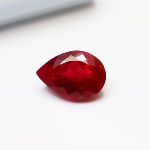 Op de afbeelding: Een gefacetteerde, peervormige robijn edelsteen. De edelsteen heeft een diepe, verzadigde rode kleur en heeft meerdere facetten die het licht reflecteren. De edelsteen staat tegen een witte achtergrond.