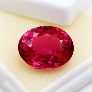 May include: A large, oval-cut, faceted ruby gemstone. The gem is a vibrant, deep red color and is set against a white background. The gemstone is displayed in a white box.