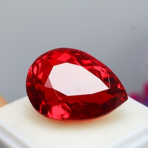 May include: A faceted, pear-shaped, red gemstone. The gem is a vibrant, deep red colour with multiple facets that reflect light. The gemstone is set on a white surface, with a soft grey background.