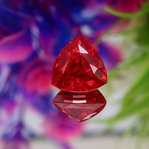 Pode incluir: Uma gema vermelha facetada em forma triangular. A gema é altamente reflexiva, com múltiplas facetas que capturam a luz. O fundo é desfocado, com cores roxas, azuis e verdes.