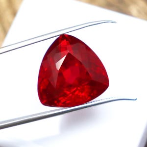 May include: A deep red triangular gemstone with a faceted cut. The gemstone is held by tweezers against a white background.