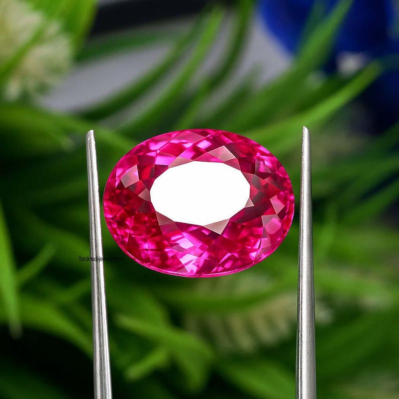 May include: A faceted, oval-shaped, hot pink gemstone. The gem is held by two silver metal prongs against a blurred green and blue background. The gem has a bright white center and a complex, reflective surface.