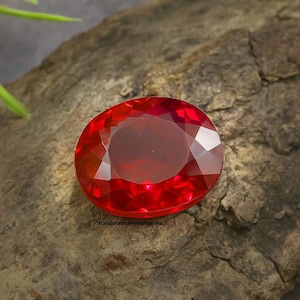 May include: A faceted, oval-shaped, red gemstone. The gem is a vibrant, deep red color and reflects light, showcasing its cut. The gemstone is set against a textured, brown surface. The text "Handmadejewelsgems" is visible.