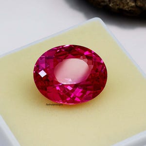 May include: A faceted, oval-shaped, hot pink gemstone. The gem is set against a light yellow background inside a white box. The text "Handmadejewelsgems" is visible on the bottom left of the gem.