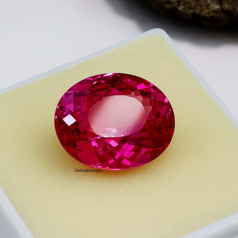 May include: A faceted, oval-shaped, hot pink gemstone. The gem is set against a light yellow background inside a white box. The text "Handmadejewelsgems" is visible on the bottom left of the gem.