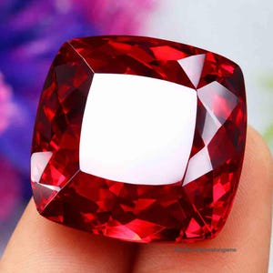 Puede incluir: Una gema grande, tallada en forma cuadrada, facetada, de color rojo intenso. La gema tiene una superficie brillante y reflectante con un área blanca central. Las facetas crean un patrón complejo de luz y sombra.