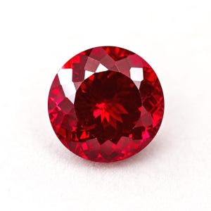 Può includere: Una pietra preziosa rotonda e sfaccettata di colore rosso intenso e vibrante. La pietra presenta un taglio a brillante con numerose sfaccettature triangolari e a forma di aquilone che riflettono la luce, creando un motivo simmetrico.