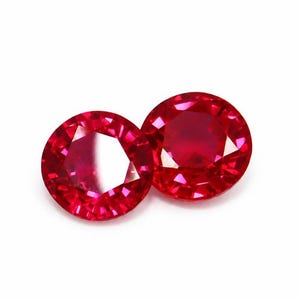 Puede incluir: Dos piedras preciosas de color rojo vibrante con corte redondo y múltiples facetas que reflejan la luz. Las piedras están colocadas una al lado de la otra, mostrando un color carmesí intenso y una superficie pulida y brillante.