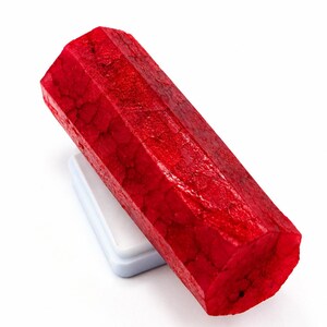 Peut inclure: Un spécimen de cristal hexagonal rouge vif avec une surface cristalline texturée. Le minéral prismatique allongé repose en biais contre une petite base rectangulaire blanche, mettant en valeur sa couleur cramoisie profonde et sa structure.
