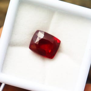 Op de afbeelding: Een dieprode, vierkant geslepen edelsteen, mogelijk een robijn, wordt getoond in een witte sieradendoos. De edelsteen heeft vele facetten die het licht reflecteren, wat een heldere schittering creëert.