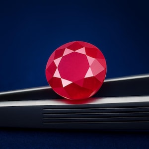 May include: A faceted, round ruby gemstone, held by dark metal tweezers. The ruby is a vibrant red colour and reflects light, showcasing its many facets. The background is a deep blue.