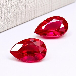 Puede incluir: Dos piedras preciosas de color rojo vibrante talladas en forma de pera con múltiples facetas que reflejan la luz. Las piedras son transparentes y muestran un tono carmesí intenso, con una superficie pulida y bordes bien definidos.
