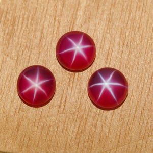 May include: Three round, cabochon-cut gemstones in a deep magenta-red color. Each stone features a distinct, centered six-rayed white star pattern, known as asterism, which radiates from the middle to the edges of the smooth, polished surface.