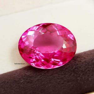 May include: A faceted, oval-shaped, hot pink gemstone. The gem has a brilliant cut, reflecting light throughout its surface. The gem is set against a neutral background. The text "Handmadejewelsngems" is visible on the lower left.