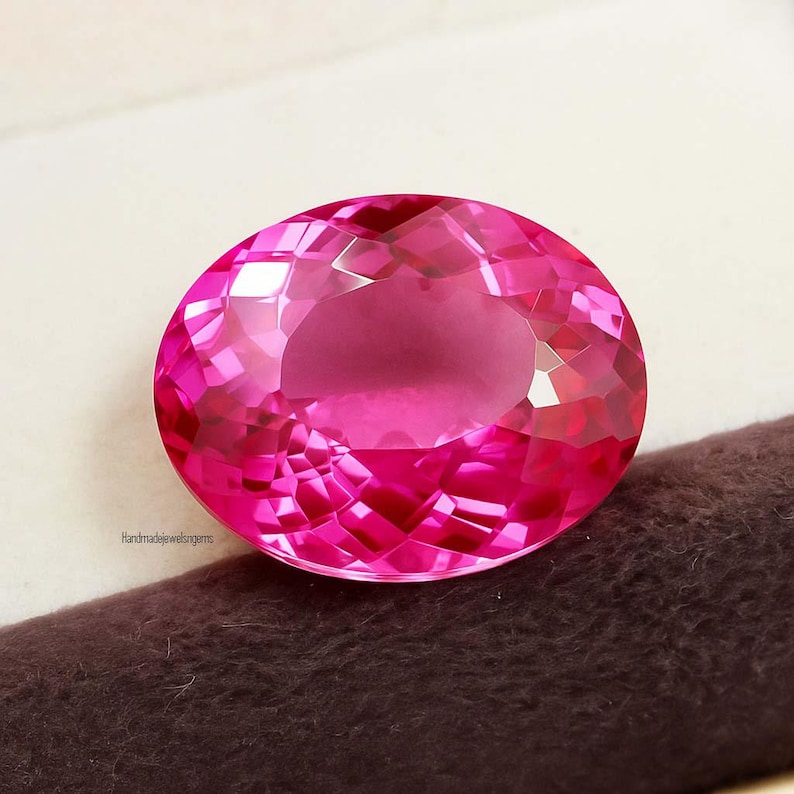 May include: A faceted, oval-shaped, hot pink gemstone. The gem has a brilliant cut, reflecting light throughout its surface. The gem is set against a neutral background. The text "Handmadejewelsngems" is visible on the lower left.
