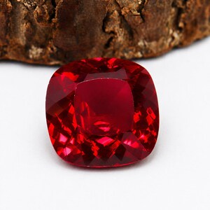 Può includere: Una gemma di rubino sfaccettata a taglio cuscino. La gemma è di un colore rosso intenso e vibrante, con molteplici sfaccettature che riflettono la luce. La gemma è posta su uno sfondo bianco, con un pezzo di legno marrone sullo sfondo.