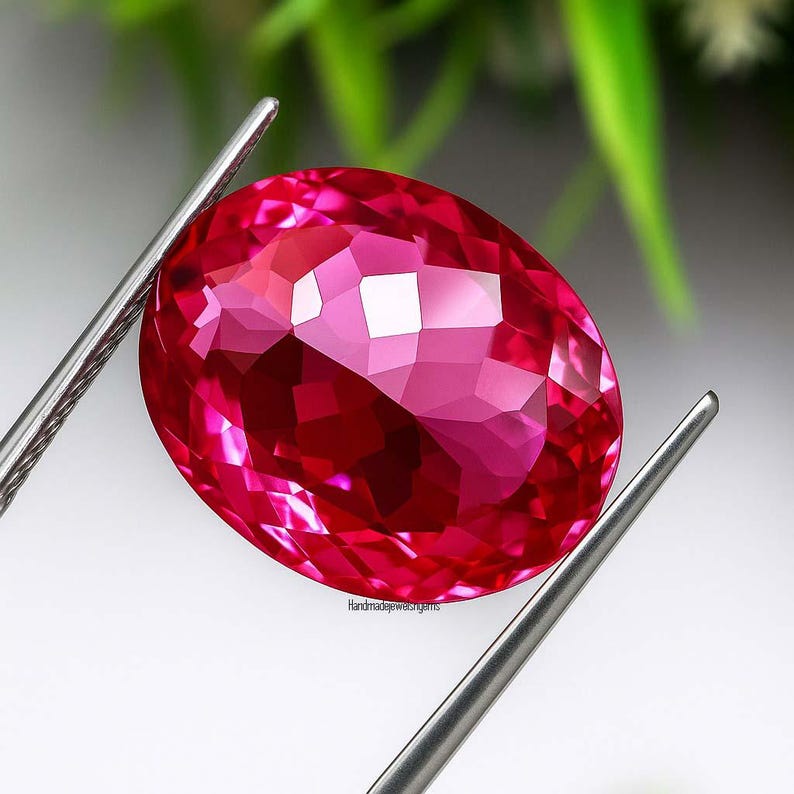 May include: A faceted, oval-cut, ruby-red gemstone is held by silver tweezers. The gem has a complex, multi-faceted surface that reflects light. The background is blurred, with hints of green foliage. The text "handmadejewelsgems" is visible.